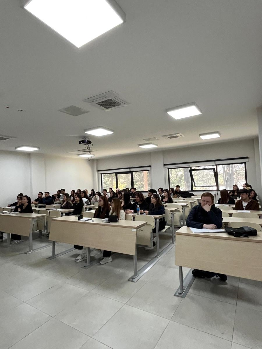 A scene from the academic advising meeting held in a modern lecture hall. The projection screen displays the title "2025-2026 Academic Year Spring Semester Civil Aviation Management Program Academic Advising Meeting." A faculty member is giving a presentation in front of the screen, accompanied by three standing students. In the foreground, students in the front rows are seen from behind, listening attentively.